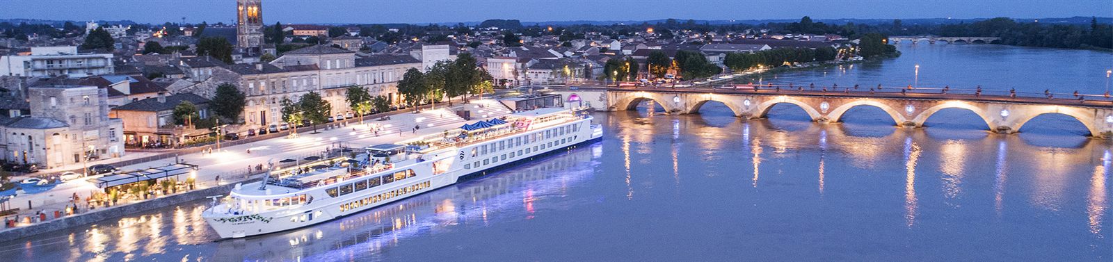 River cruise ship lit at dusk by a stone bridge